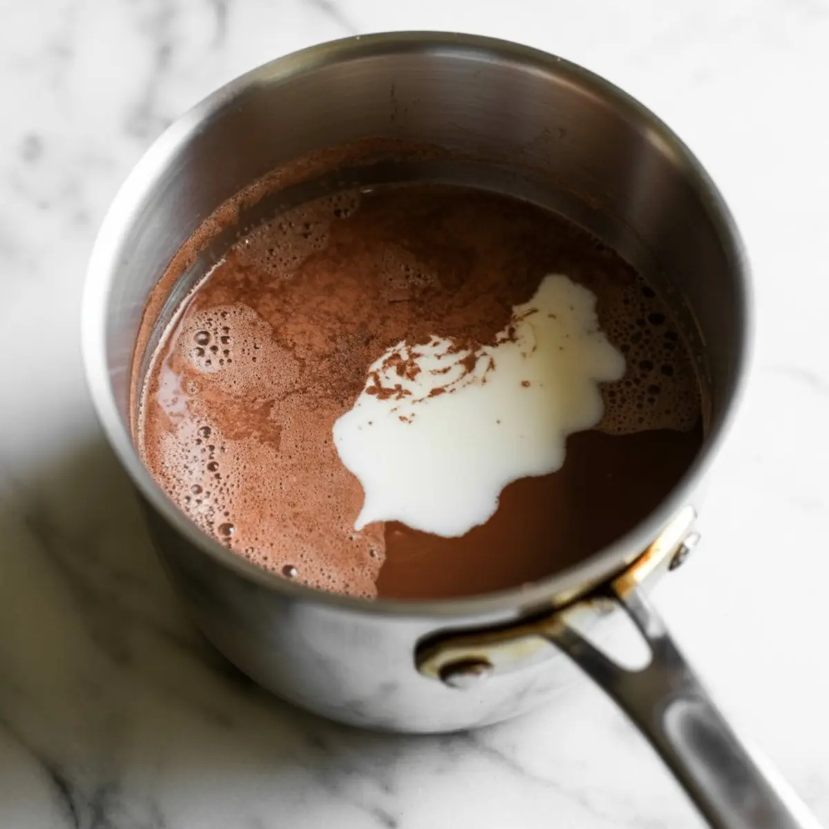 Steel saucepan with a chocolate mixture and a swirl of cream on a white marble surface, showing the beginning stage of making frozen hot chocolate.