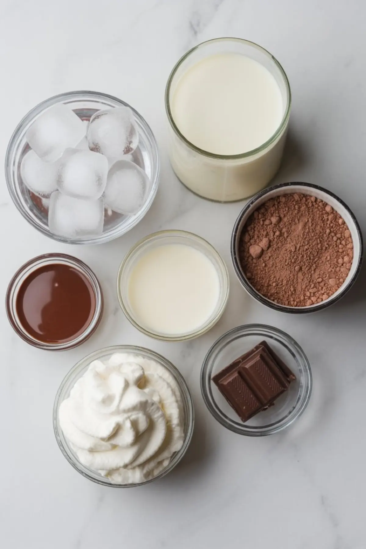 Flat lay of ingredients for frozen hot chocolate including ice cubes, milk, cocoa powder, chocolate syrup, whipped cream, chocolate squares, and sweetened condensed milk in glass bowls.