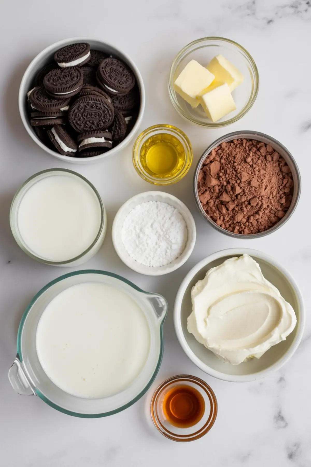 An overhead flat lay arranges Oreo cookies, cocoa powder, powdered sugar, cream cheese, milk, heavy cream, butter cubes, vanilla, and oil in small bowls, capturing organized ingredients for chocolate dirt cake.
