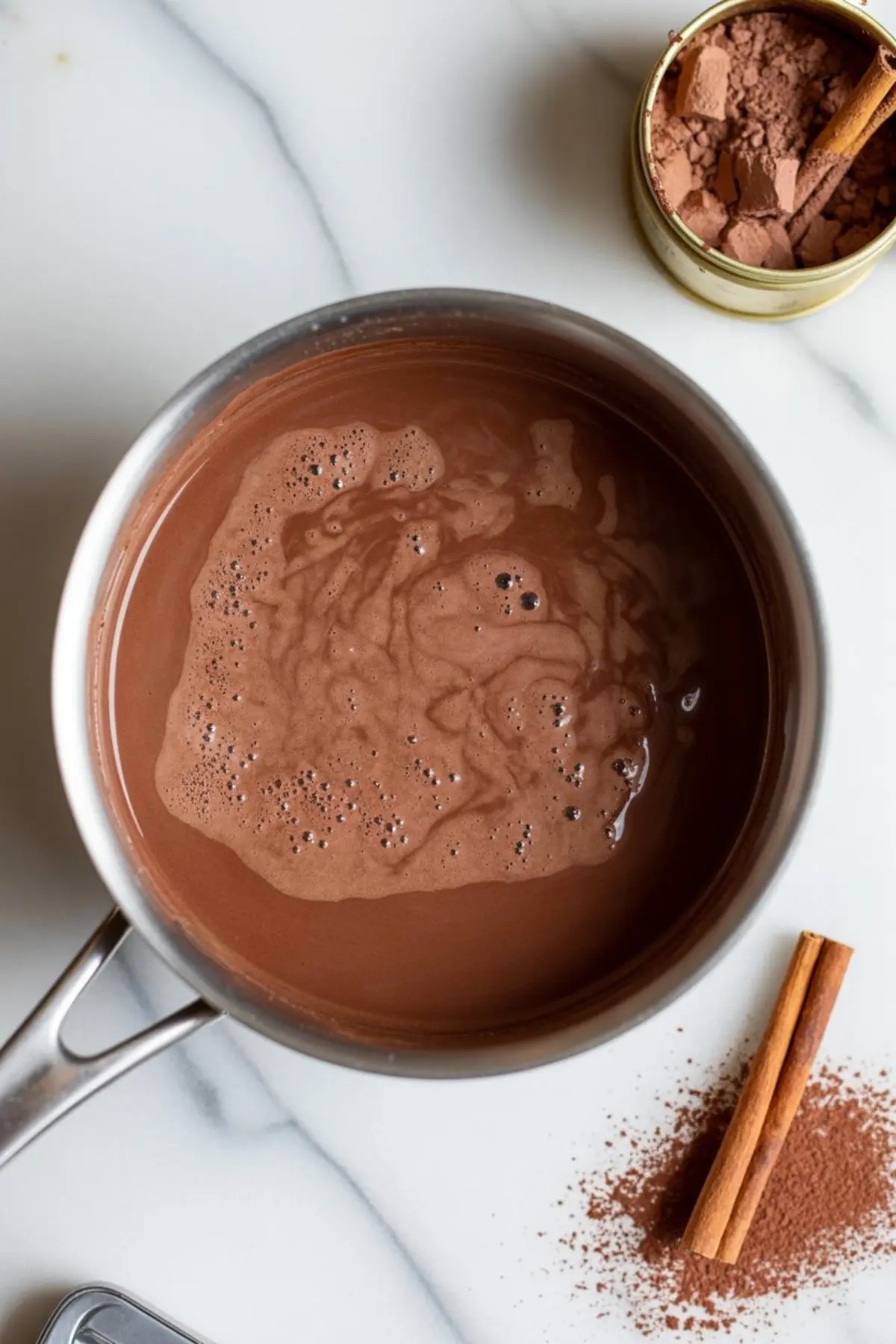 Stainless steel saucepan filled with milk, dark chocolate chunks, and cocoa powder ready to be whisked on a white countertop.
