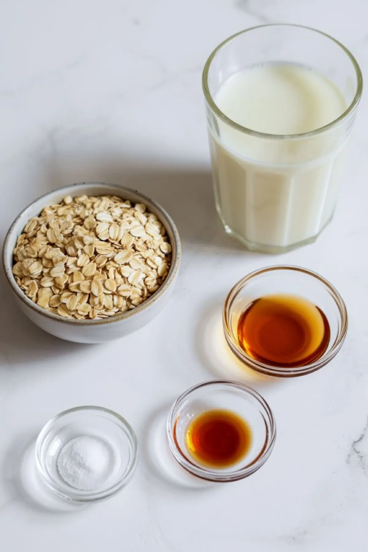 Flat lay of oat milk ingredients on a white countertop, including a bowl of rolled oats, a glass of plant-based milk, and small bowls with vanilla extract, maple syrup, and salt.