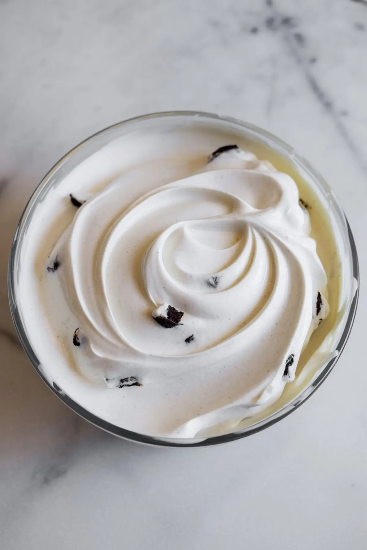 Swirled whipped cream topping with crushed cookie bits in a glass bowl, likely part of a layered dessert or cake filling, placed on a marble counter.
