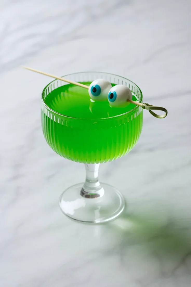 Bright green Halloween cocktail in a vintage coupe glass, garnished with a skewer of candy eyeballs for a spooky drink presentation.