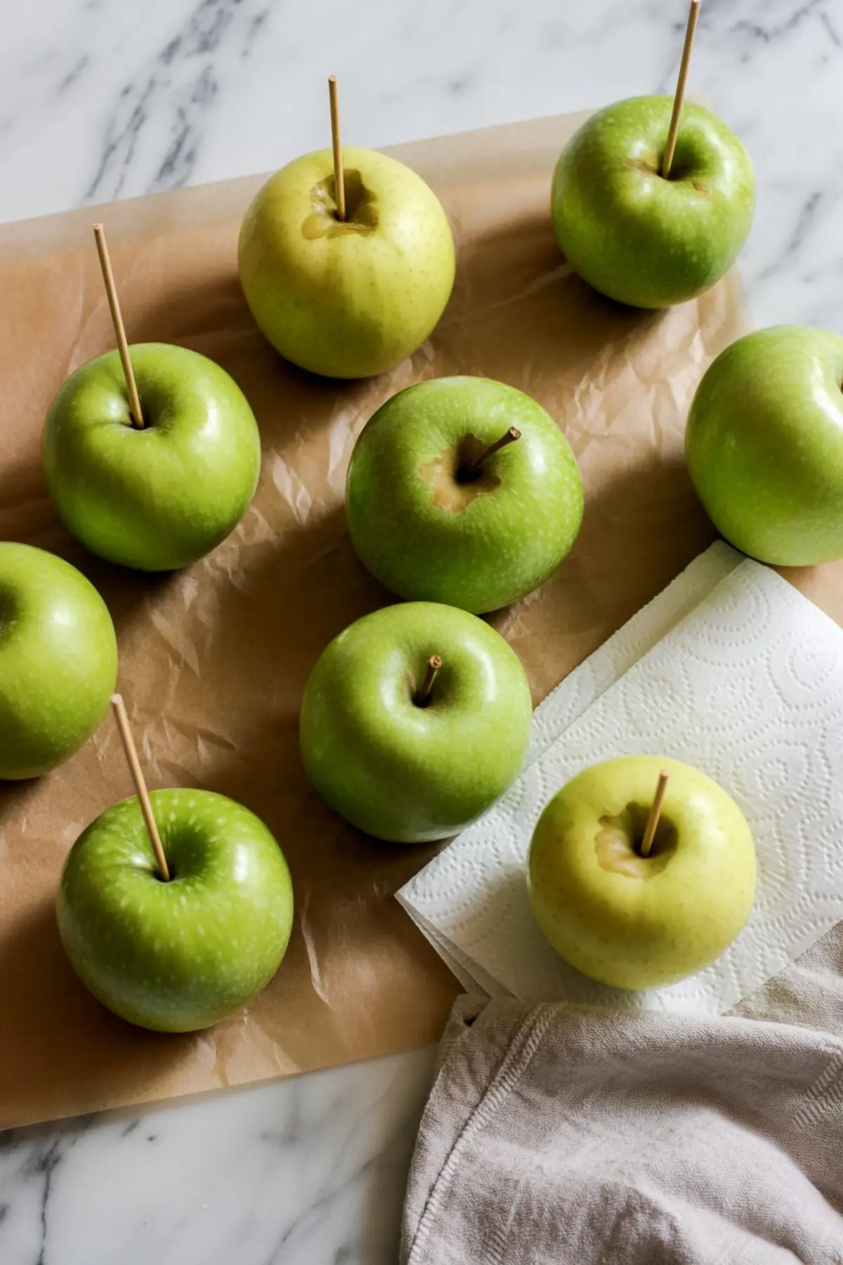 Green apples with wooden sticks inserted, arranged on parchment paper over a marble countertop, prepped for caramel apple dipping.