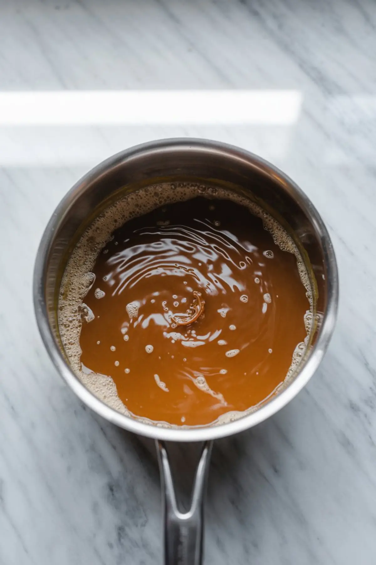 Pot of golden brown caramel sauce swirled in a stainless steel saucepan, freshly cooked and ready for dipping apples.