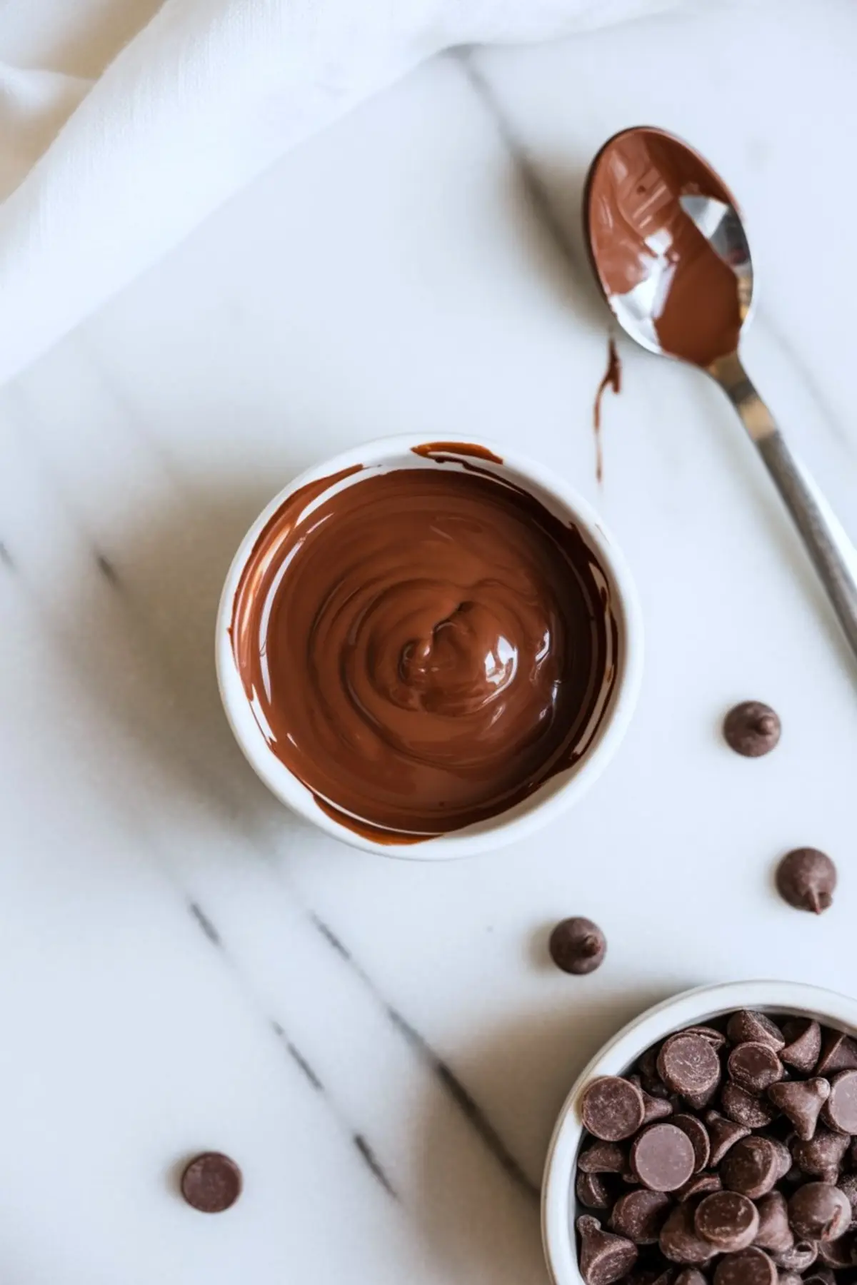 Small white bowl filled with melted chocolate on a marble surface, surrounded by chocolate chips, a silver spoon, and a white cloth napkin.
