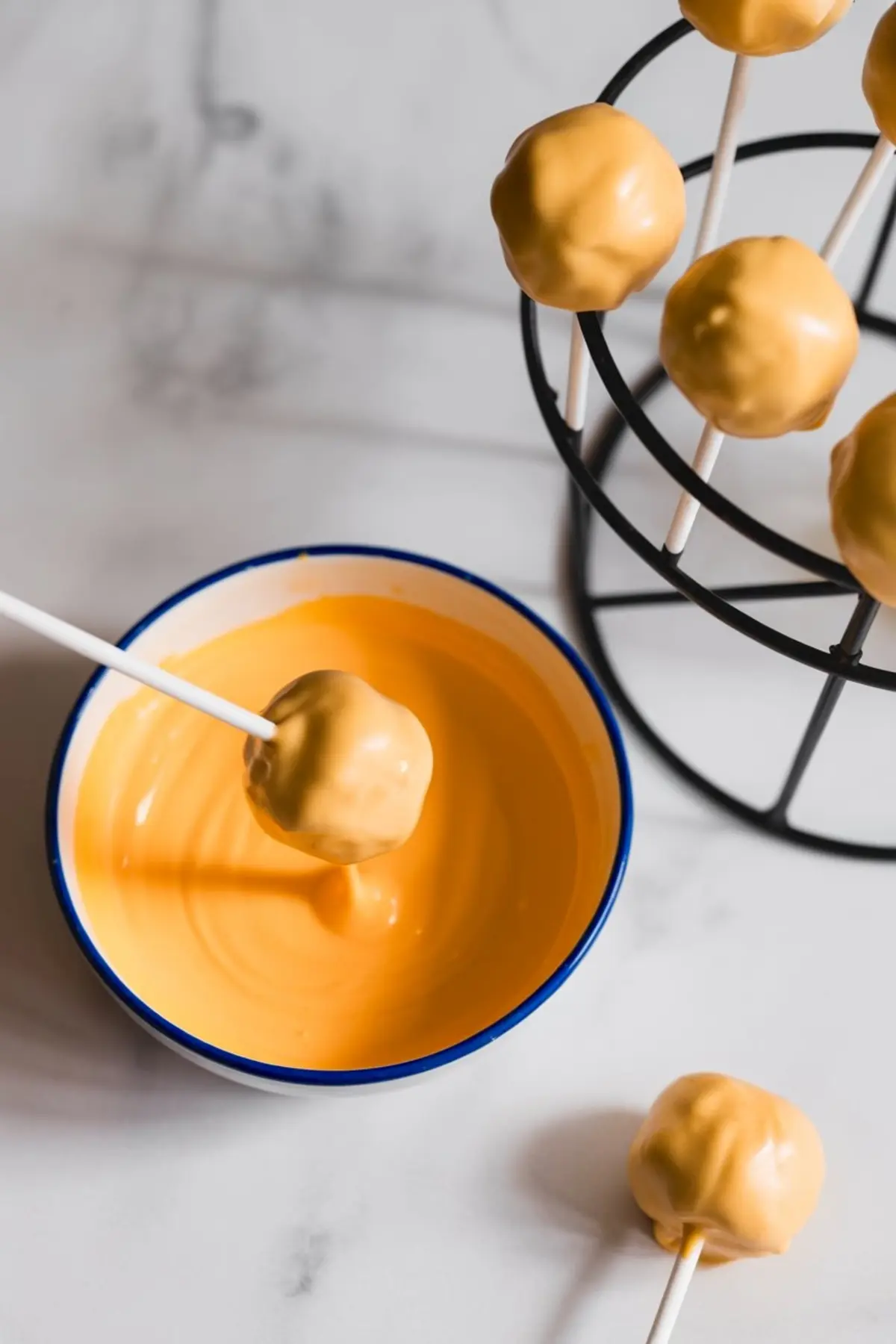 Cake pop being dipped into a bowl of smooth orange candy melt with several coated cake pops setting on a metal stand in the background.
