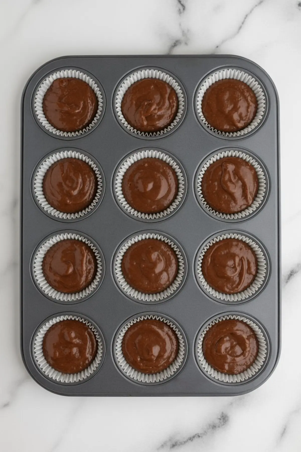 Metal muffin pan lined with cupcake papers, each filled with chocolate batter, ready for baking.