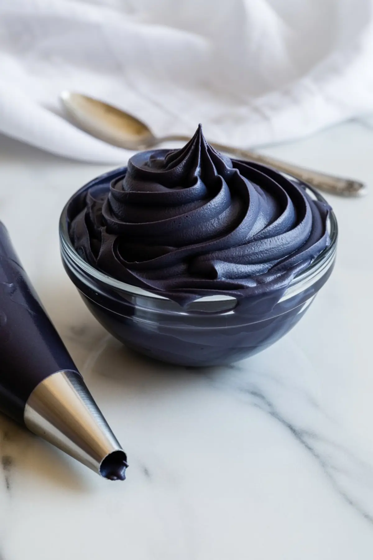Glass bowl of glossy black buttercream frosting with a piping bag beside it on a marble surface.