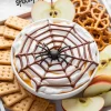 Halloween dessert dip topped with whipped cream, caramel drizzle, and a chocolate spiderweb design, surrounded by apple slices, pretzels, and crackers.