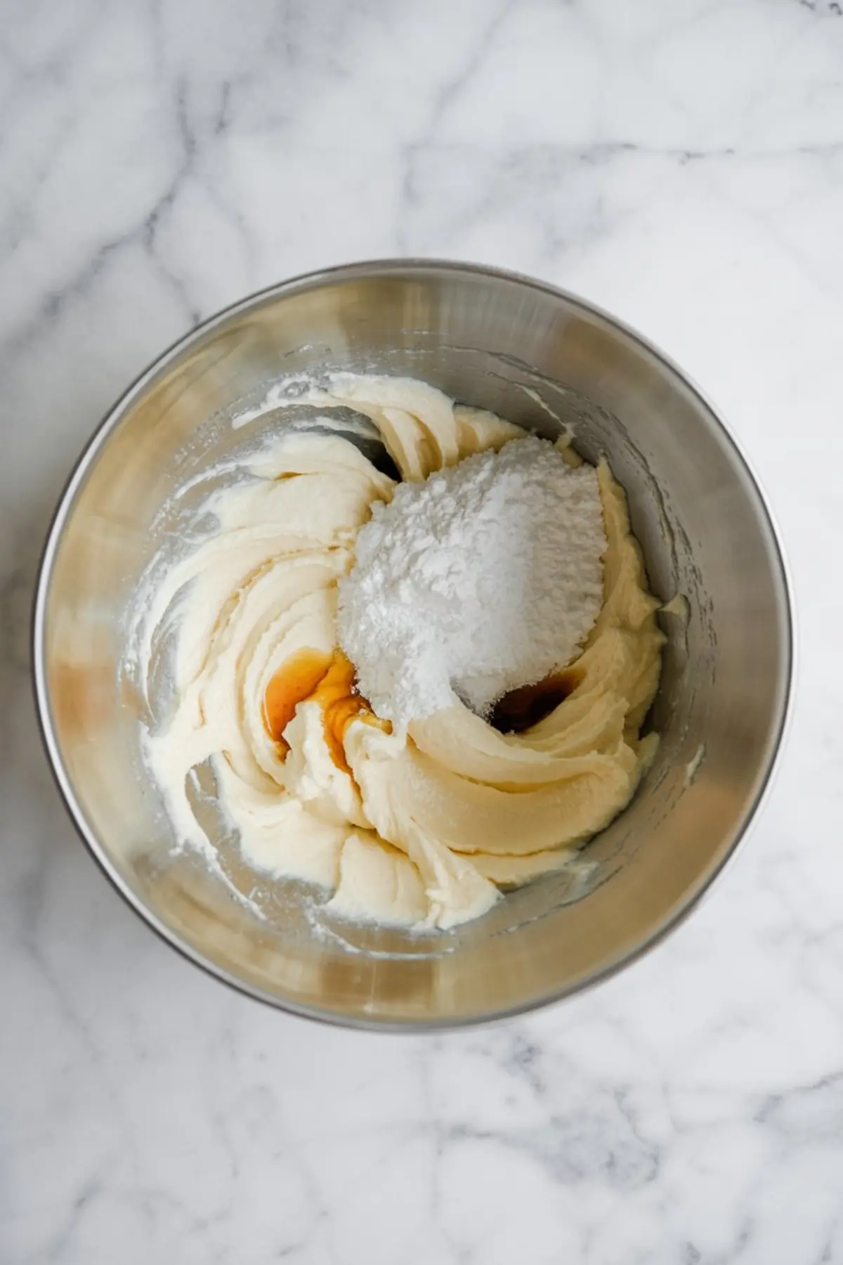 Stainless steel mixing bowl with creamed butter, powdered sugar, and vanilla extract on a white marble surface.