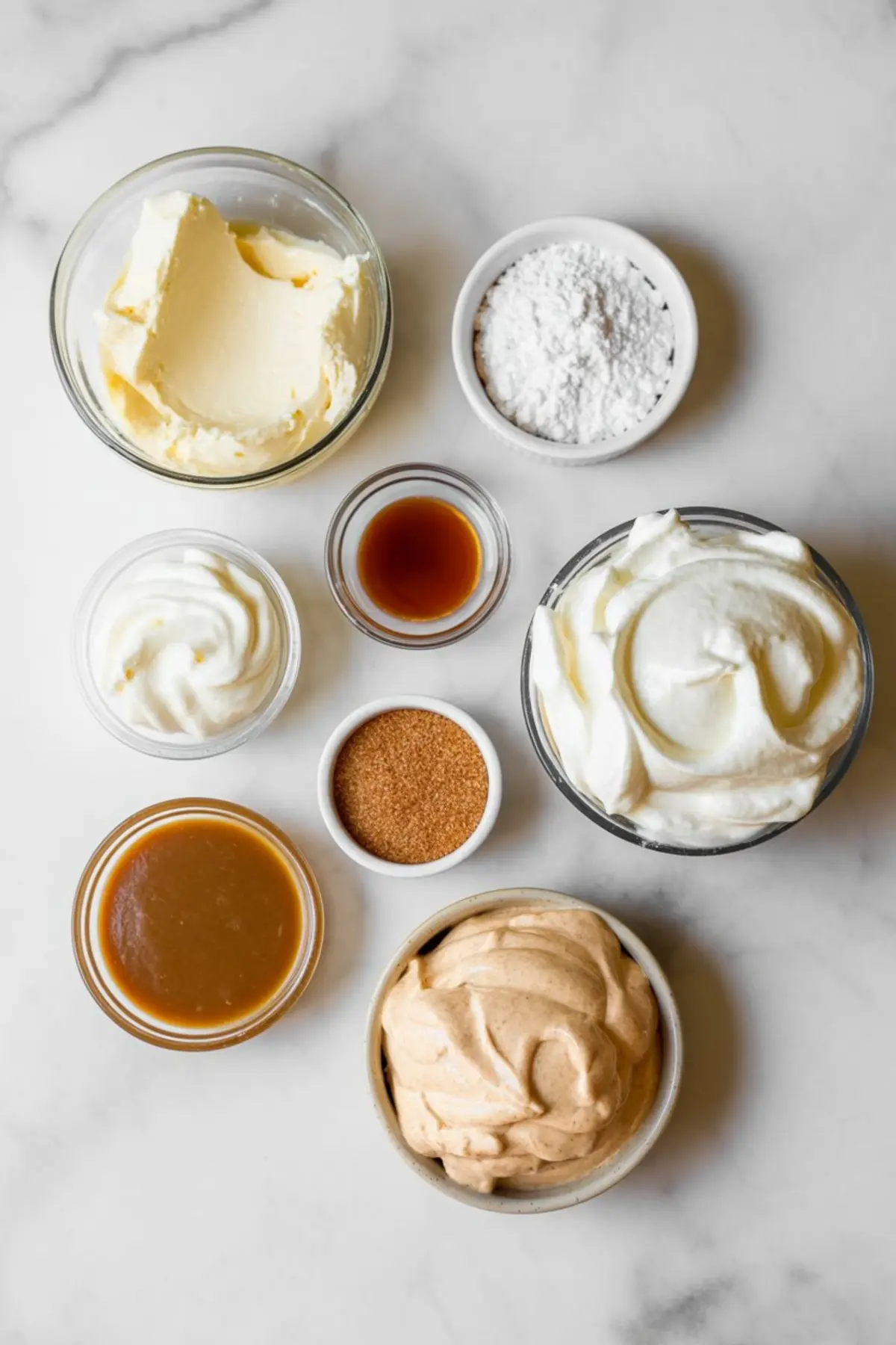 Overhead view of small bowls filled with baking ingredients including butter, powdered sugar, vanilla extract, yogurt, caramel sauce, brown sugar, whipped cream, and spiced cream mixture.