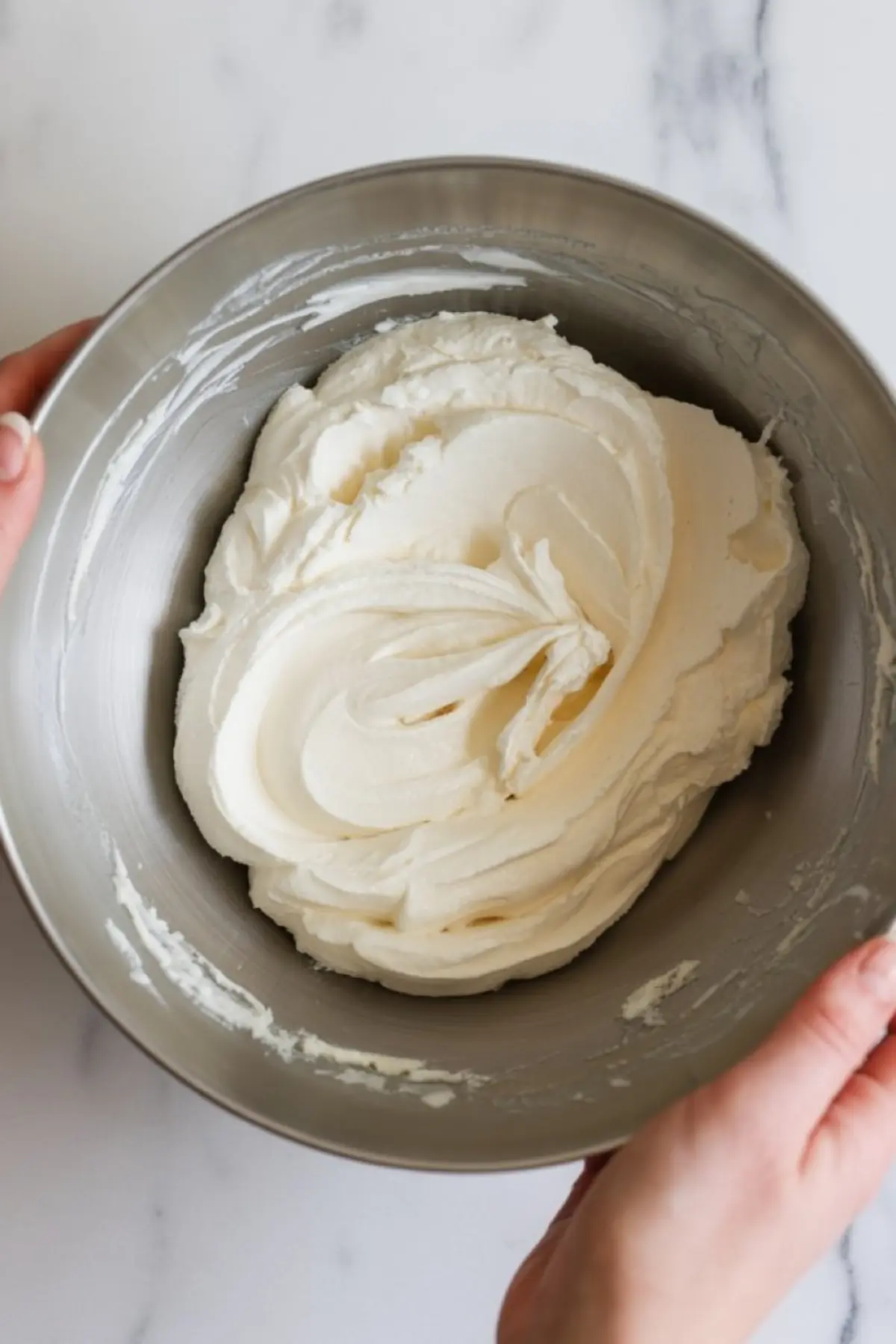 Hands holding a metal mixing bowl filled with smooth, freshly whipped cream frosting.