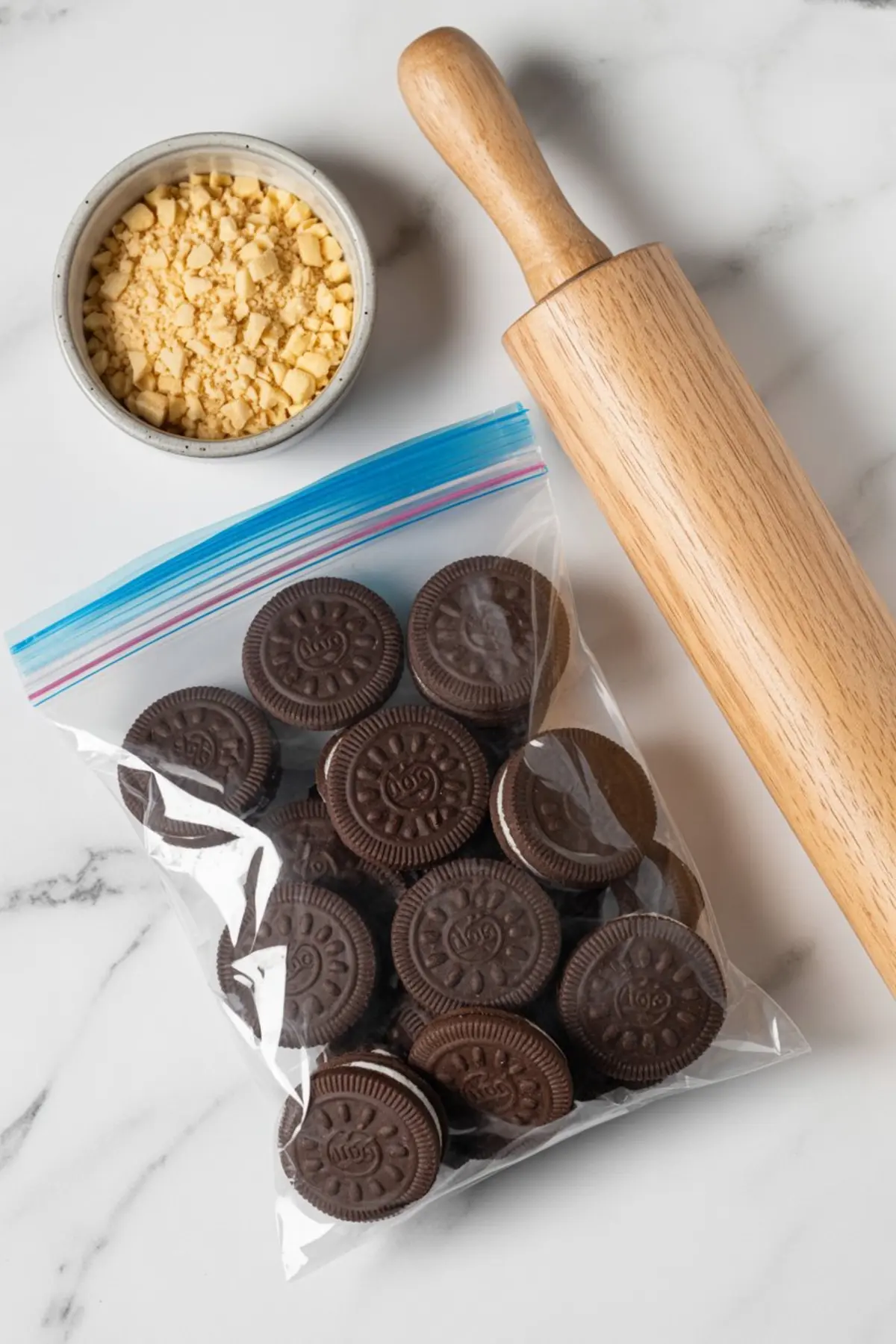 Flat lay of whole chocolate sandwich cookies in a clear zip-top bag, a bowl of crushed cookies, and a wooden rolling pin on a white marble surface, showing ingredients and tools for making crushed cookie bases.
