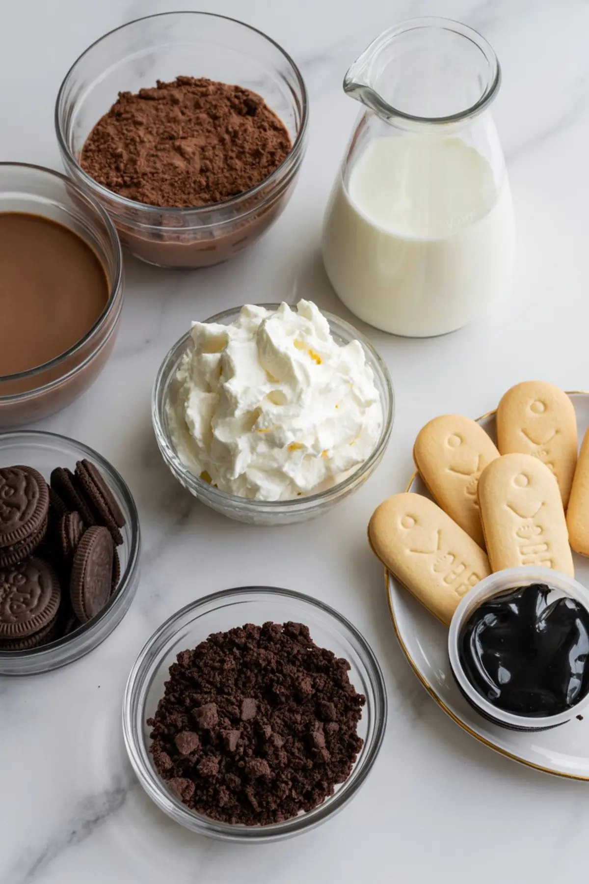 Assorted dessert ingredients on a white marble counter including chocolate pudding, cocoa powder, whipped cream, chocolate sandwich cookies, crushed chocolate cookies, vanilla cookies, black gel icing, and a glass jug of milk.
