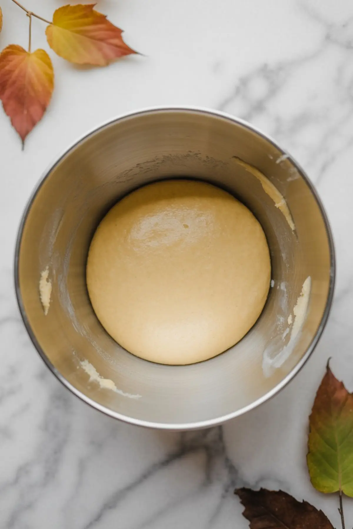 Smooth, risen dough resting in a metal mixing bowl, surrounded by scattered autumn leaves on a white marble surface.
