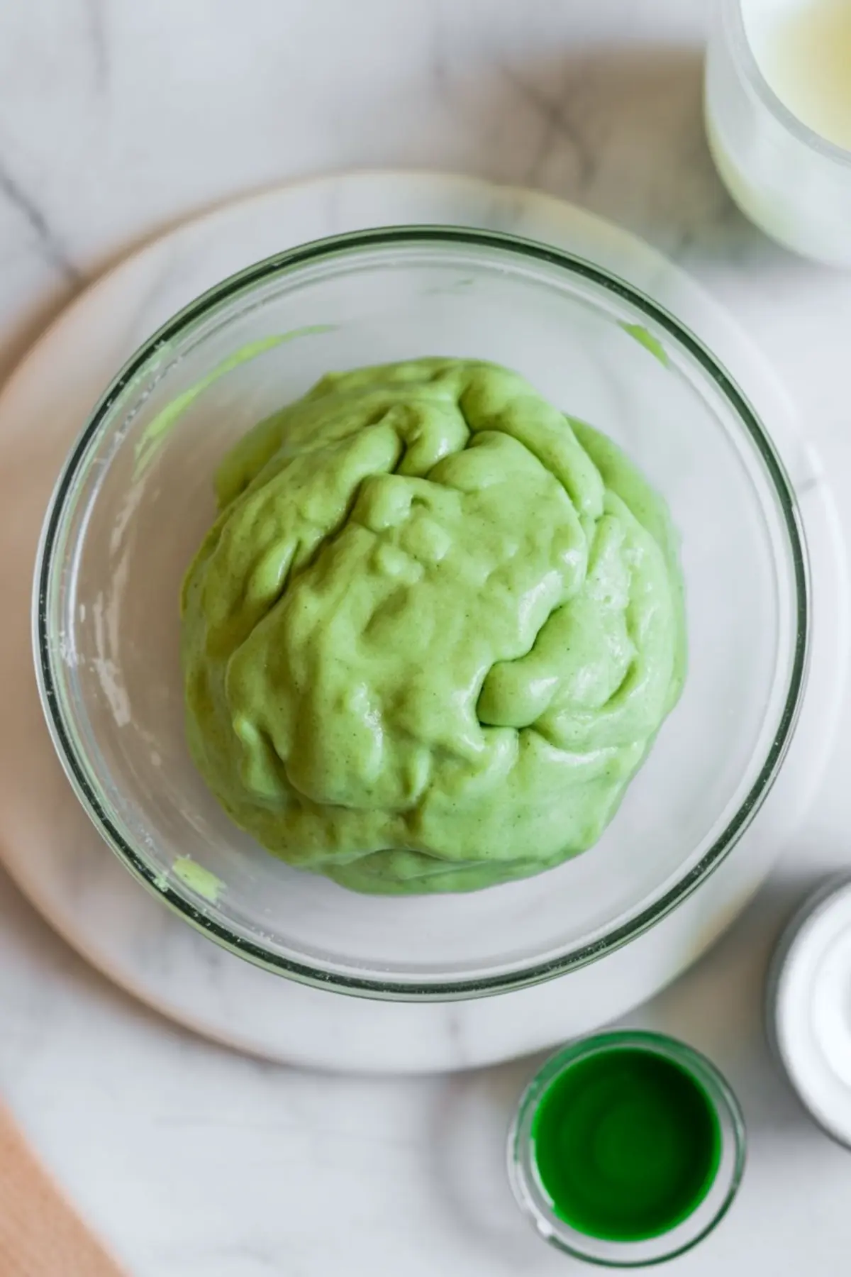 Bright green cookie dough formed into a ball in a clear glass bowl, placed beside a small bowl of vibrant green food coloring on a light marble background.
