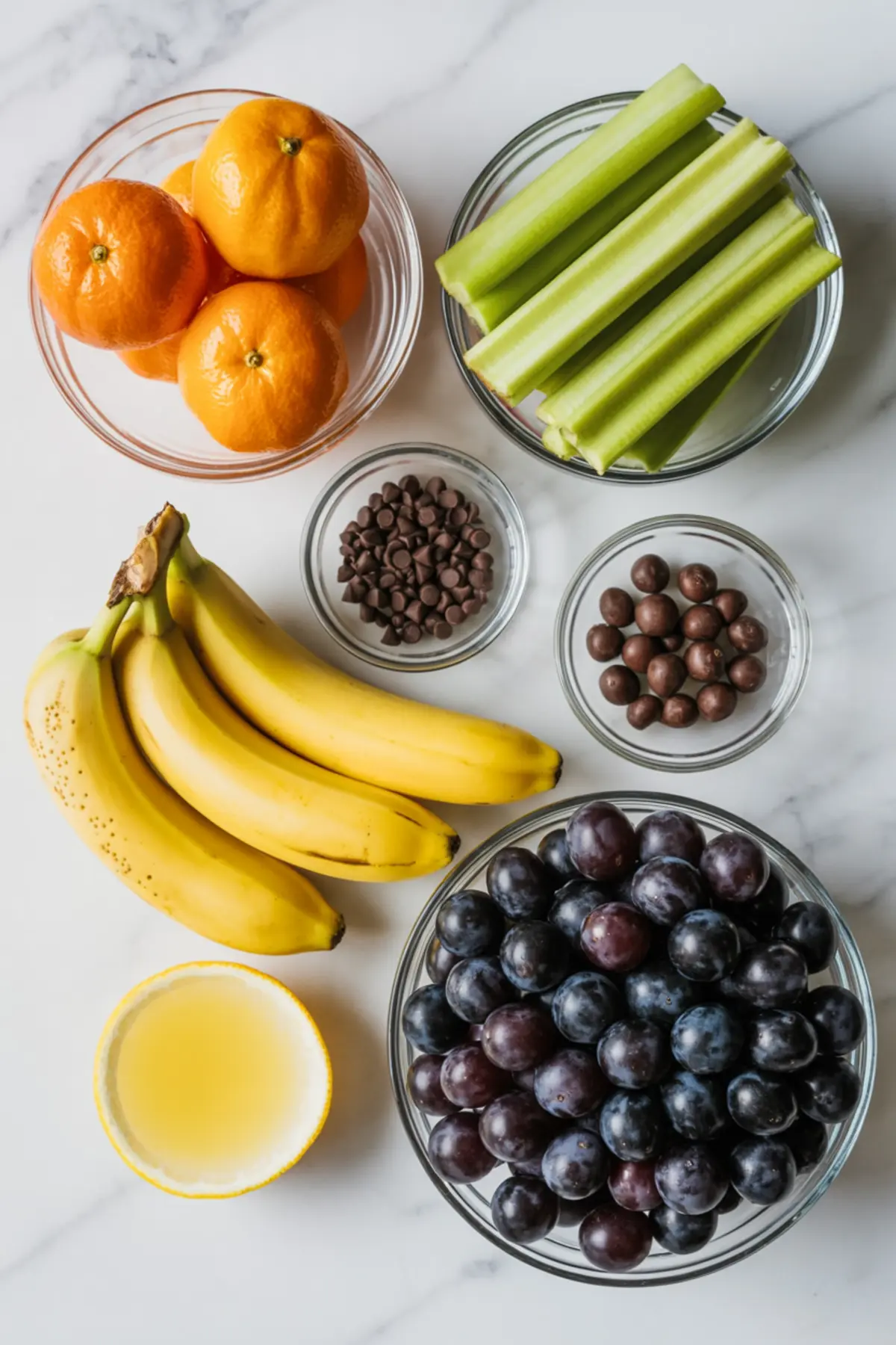 Overhead view of ingredients for Halloween fruit snacks including bananas, peeled mandarins, celery sticks, black grapes, chocolate chips, malt balls, and a halved lemon on a white marble surface.