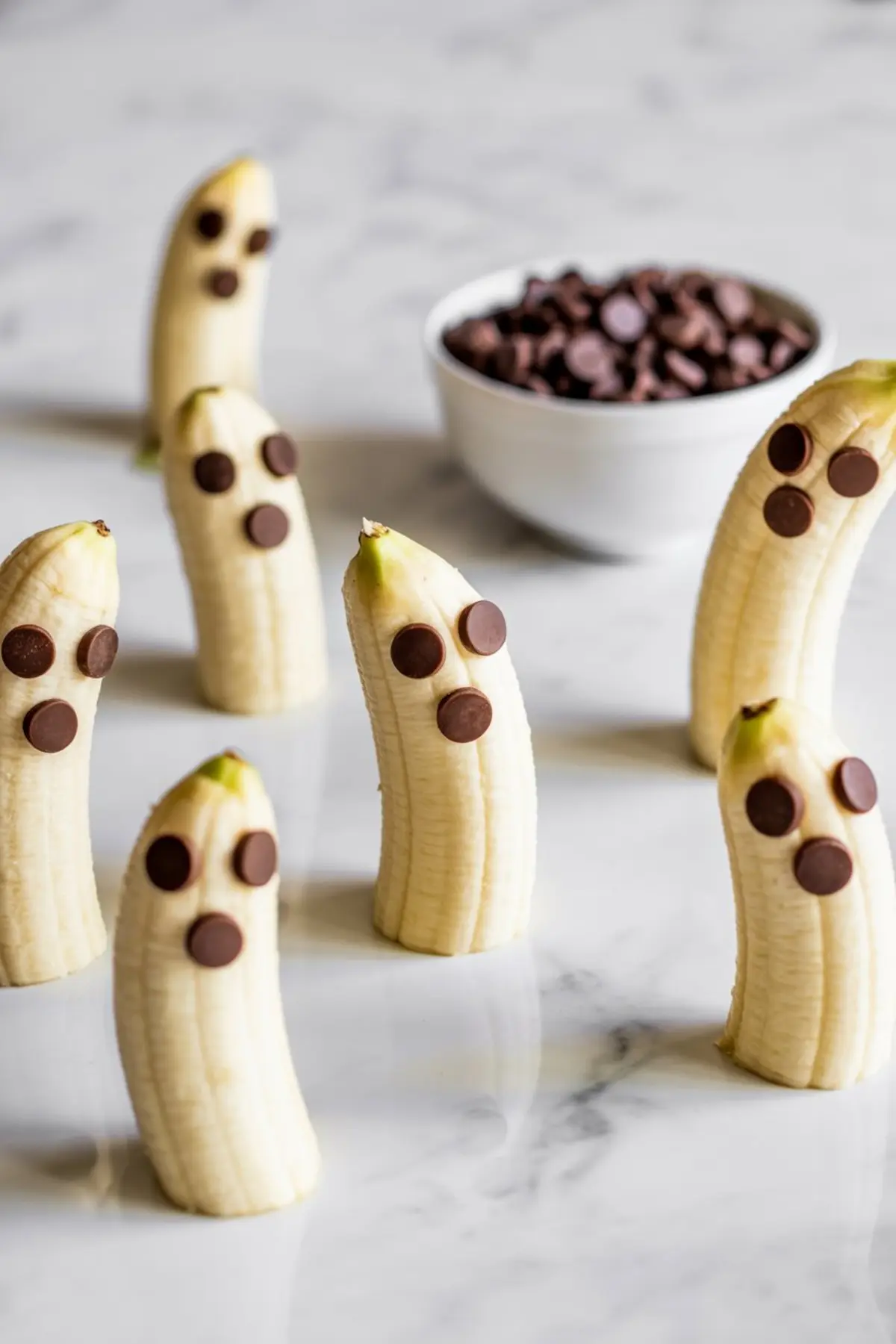 Group of bananas cut in half and decorated with chocolate chips to resemble spooky Halloween ghosts, arranged on a white marble background with a bowl of chocolate chips.