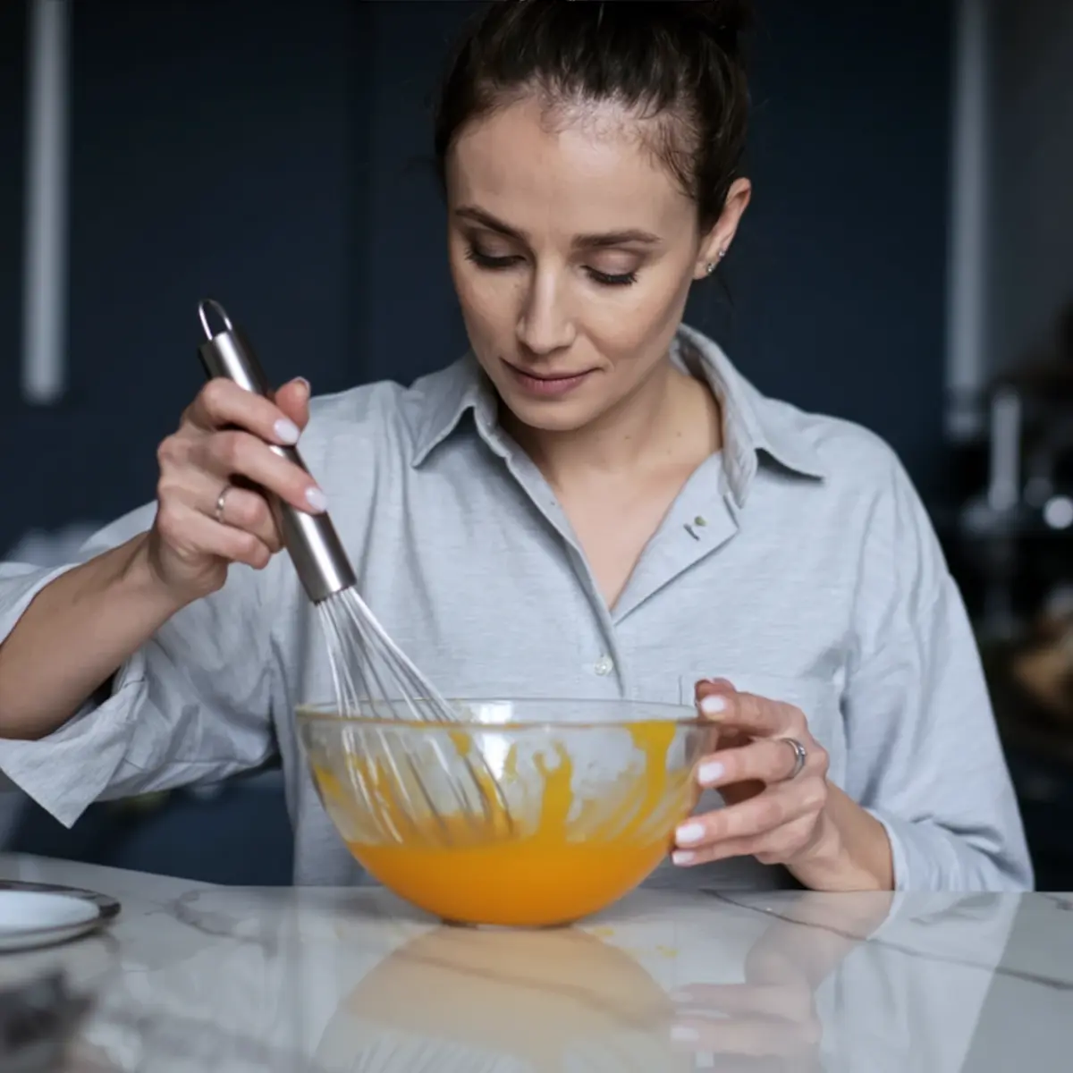 Emma whisking orange-colored macaron batter in a clear glass mixing bowl while standing at a marble countertop in a modern kitchen.