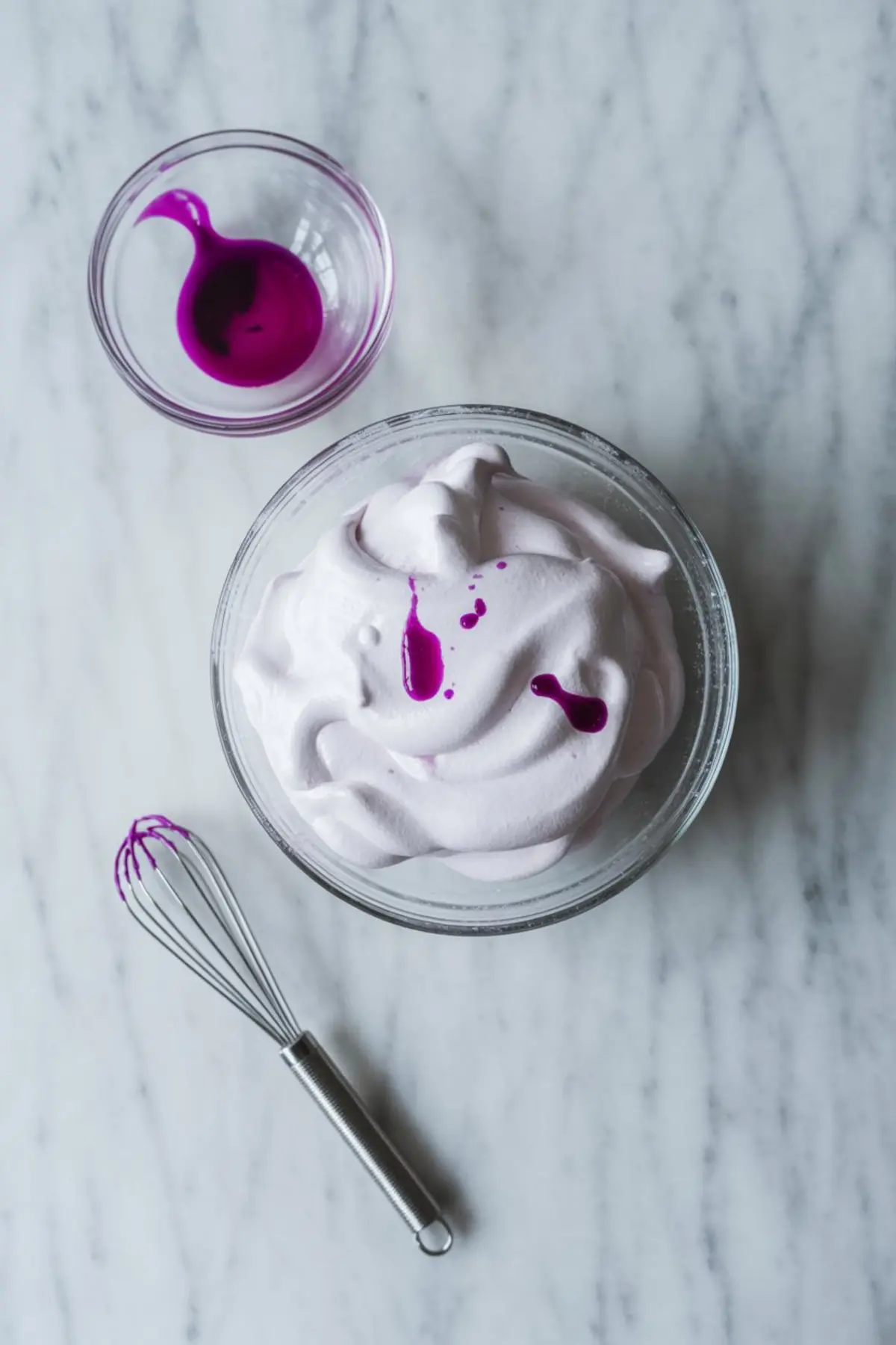 Bowl of pale purple meringue with drops of vibrant purple gel food coloring on top, placed beside a whisk and a small glass dish of dye.
