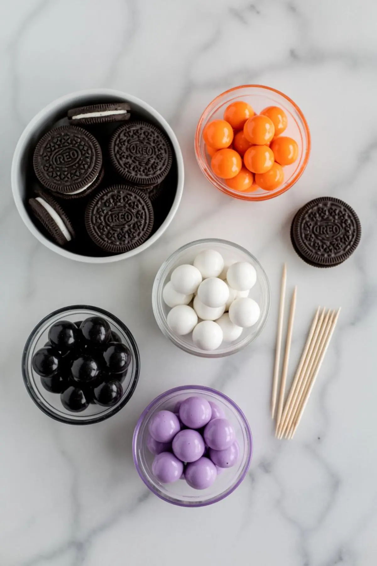 Flat lay of Halloween treat ingredients on a white marble surface, including Oreo cookies, orange, white, black, and purple candy coating balls in small bowls, and wooden toothpicks.