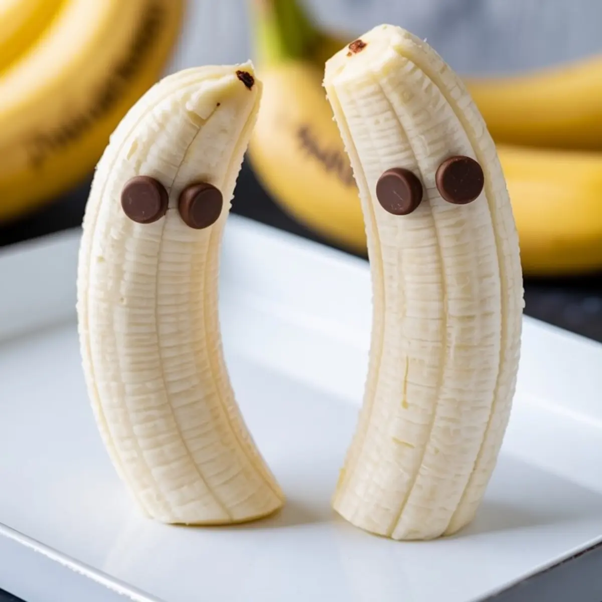 Two peeled bananas standing upright with chocolate chips pressed into them to resemble ghost faces, placed on a white tray with fresh bananas in the background.