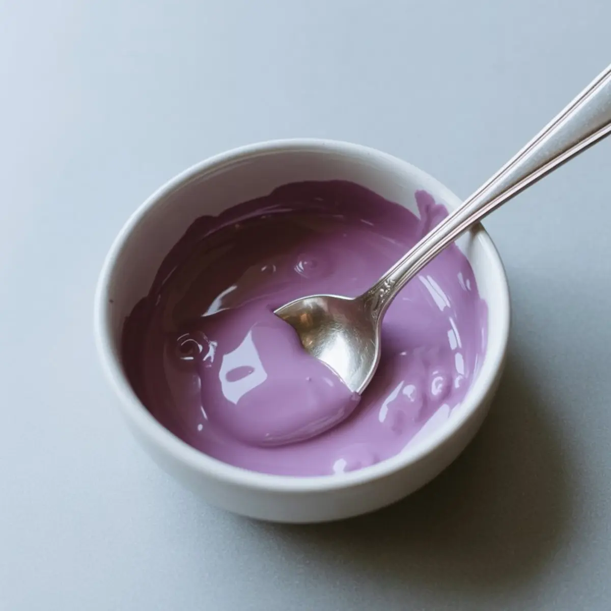 Small white bowl filled with thick purple yogurt or pudding, with a silver spoon resting inside, placed on a smooth light gray surface.