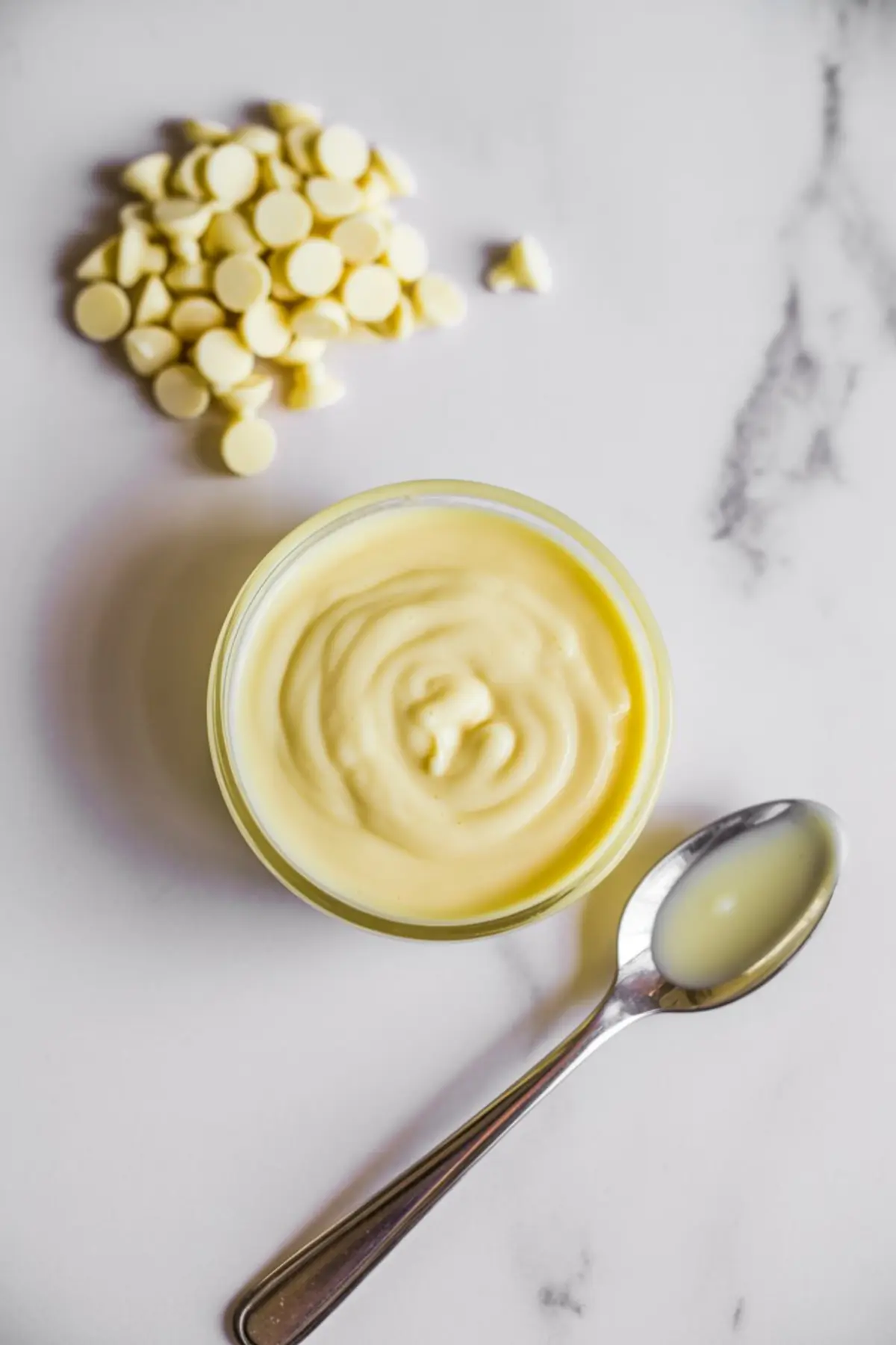 Melted white chocolate in a glass bowl with a metal spoon, set beside a small mound of white chocolate chips on a white marble surface.