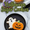 Decorated Halloween sugar cookies shaped like a ghost and jack-o’-lantern on a black plate, with additional purple, black, and orange cookies on a tray in the background.