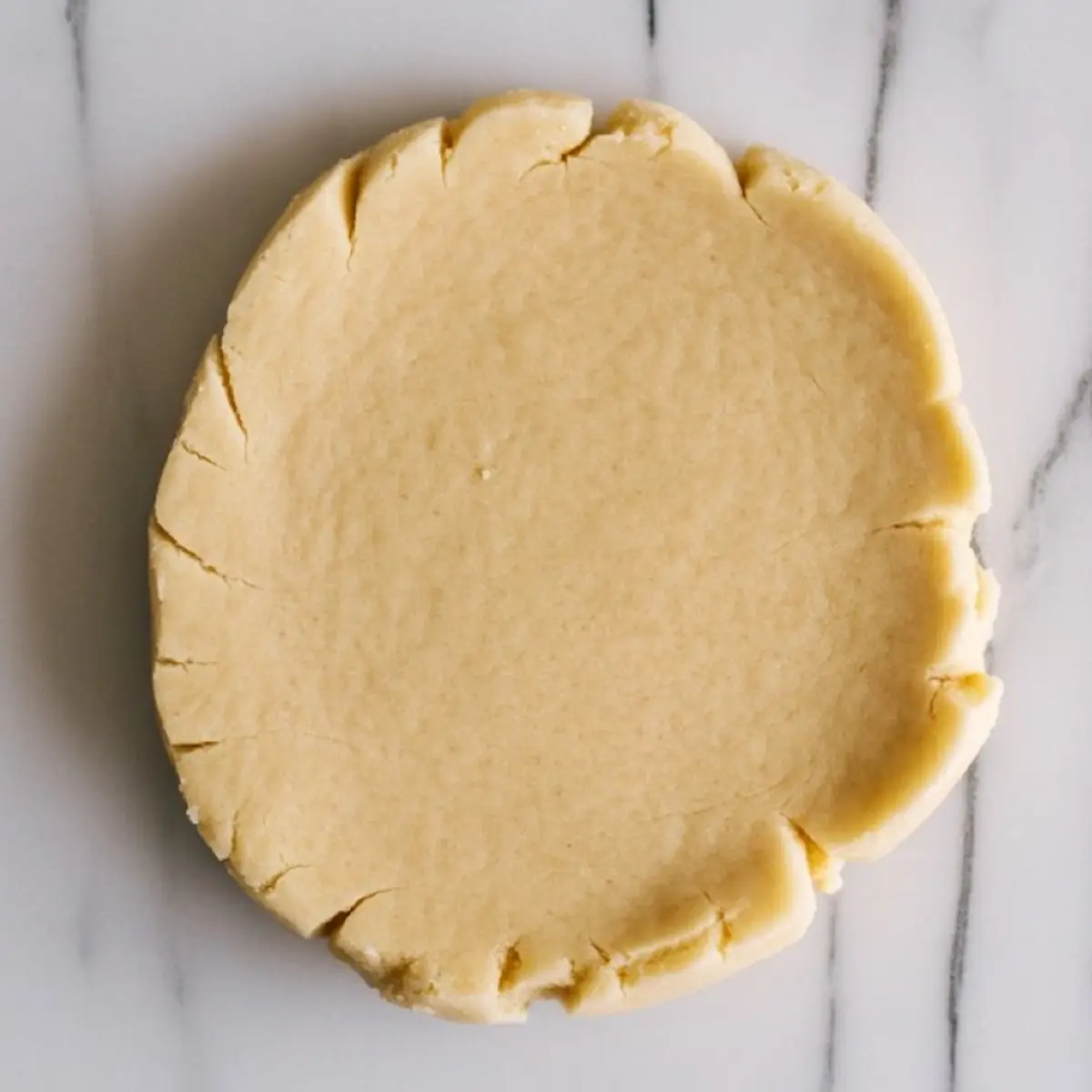 Round disc of sugar cookie dough with cracked edges resting on a marble surface, prepared for rolling and cutting.
