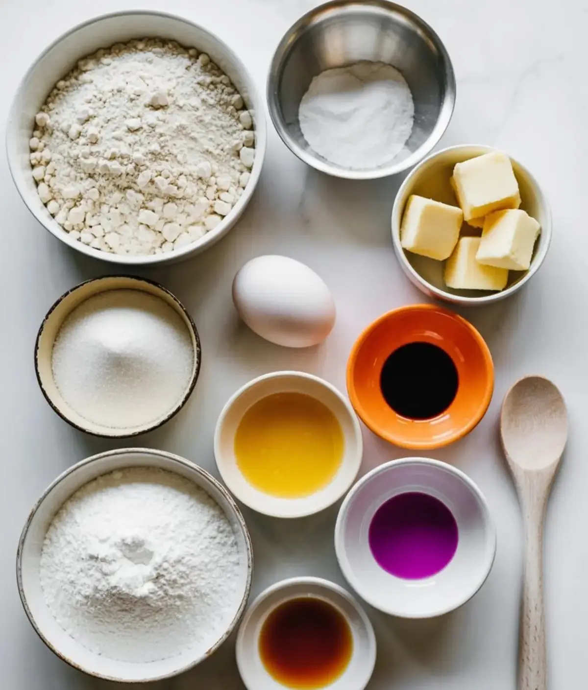 Flat lay of sugar cookie ingredients including flour, powdered sugar, butter, egg, food coloring, vanilla extract, and baking powder arranged in bowls on a white surface.
