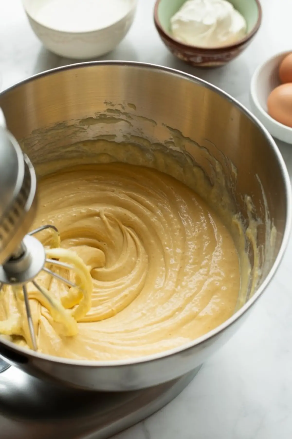 Mixing bowl with creamy yellow cake batter being whipped in a stand mixer, with eggs, sour cream, and milk in bowls nearby on a marble countertop.