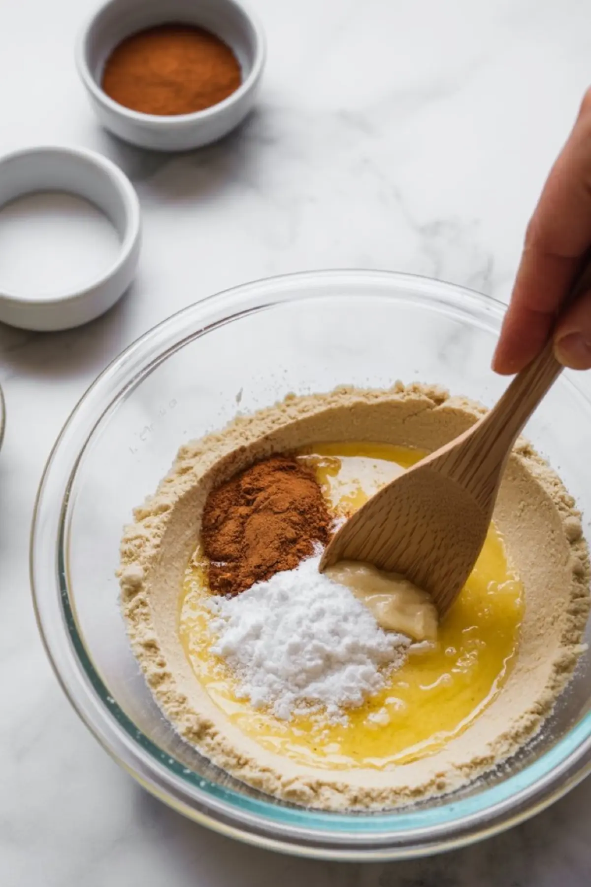 Wooden spoon mixing flour, baking powder, cinnamon, mashed banana, and melted butter in a clear bowl for baking preparation.
