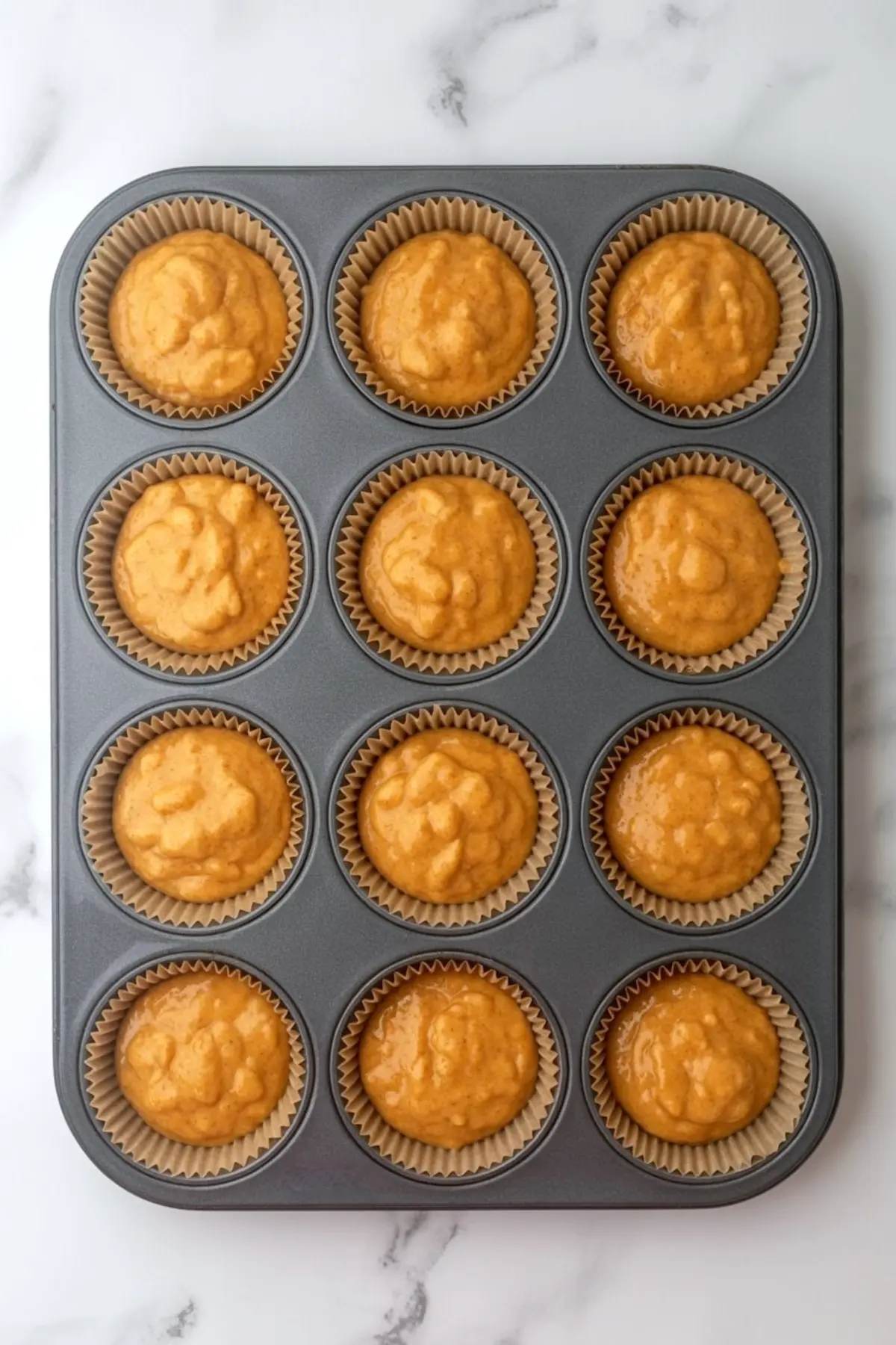 Muffin tin filled with unbaked keto pumpkin muffin batter in paper liners on a marble surface.