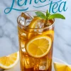 Close-up of iced lemon tea in a tall glass with lemon slices, ice cubes, and mint garnish, with “Lemon tea” text in cursive above on a bright marble tabletop.