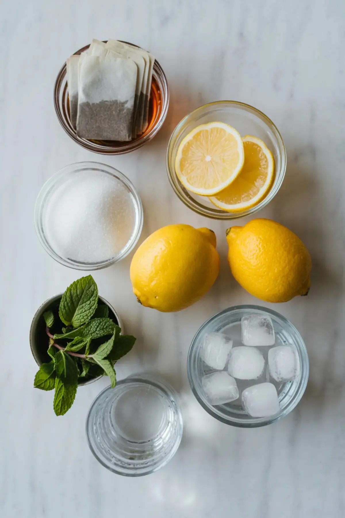 Flat lay of lemon tea ingredients including tea bags, lemon slices, whole lemons, fresh mint, ice cubes, sugar, and water on a white marble countertop.
