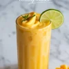 Frozen mango smoothie in a tall glass with a thick swirl top, garnished with fresh mint and lime slice, set on a marble surface next to cubed mango, labeled as a healthy drink option.