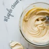Whipped maple cream cheese frosting in a glass mixing bowl with electric beaters, next to a frosting spreader coated with a swirl of creamy frosting on a marble surface.