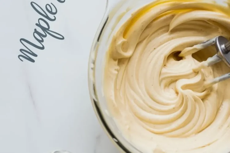 Whipped maple cream cheese frosting in a glass mixing bowl with electric beaters, next to a frosting spreader coated with a swirl of creamy frosting on a marble surface.