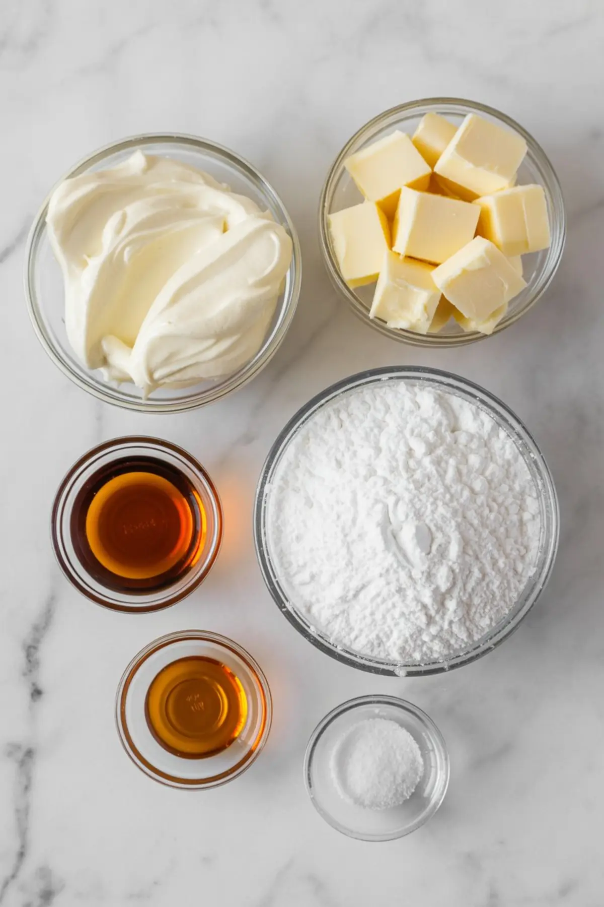 Flat lay of maple cream cheese frosting ingredients in glass bowls, including butter cubes, cream cheese, powdered sugar, vanilla extract, maple syrup, and a pinch of salt.