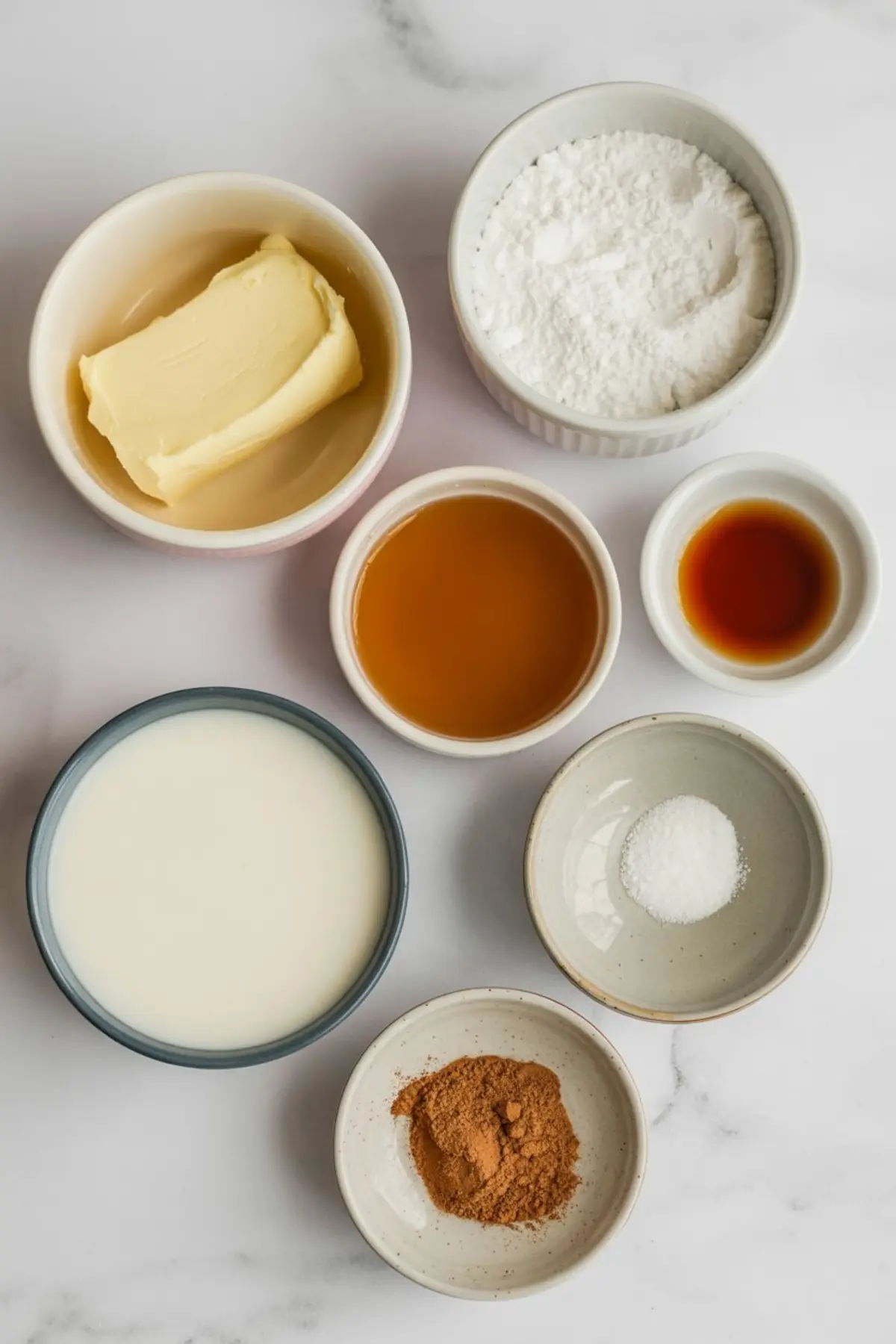 Flat lay of ingredients for maple frosting including butter, powdered sugar, maple syrup, vanilla extract, milk, cinnamon, and salt in separate ceramic bowls on a marble background.
