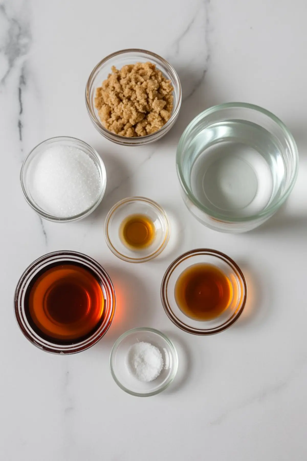 Flat lay of maple syrup ingredients in small glass bowls, including brown sugar, white sugar, water, vanilla extract, maple extract, corn syrup, and salt on a white marble surface.