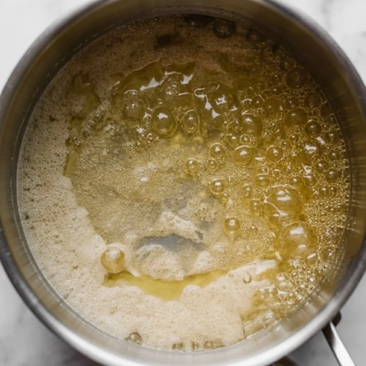 Close-up of bubbling homemade syrup mixture cooking in a saucepan, highlighting the golden brown color and thickening texture during the simmering stage.