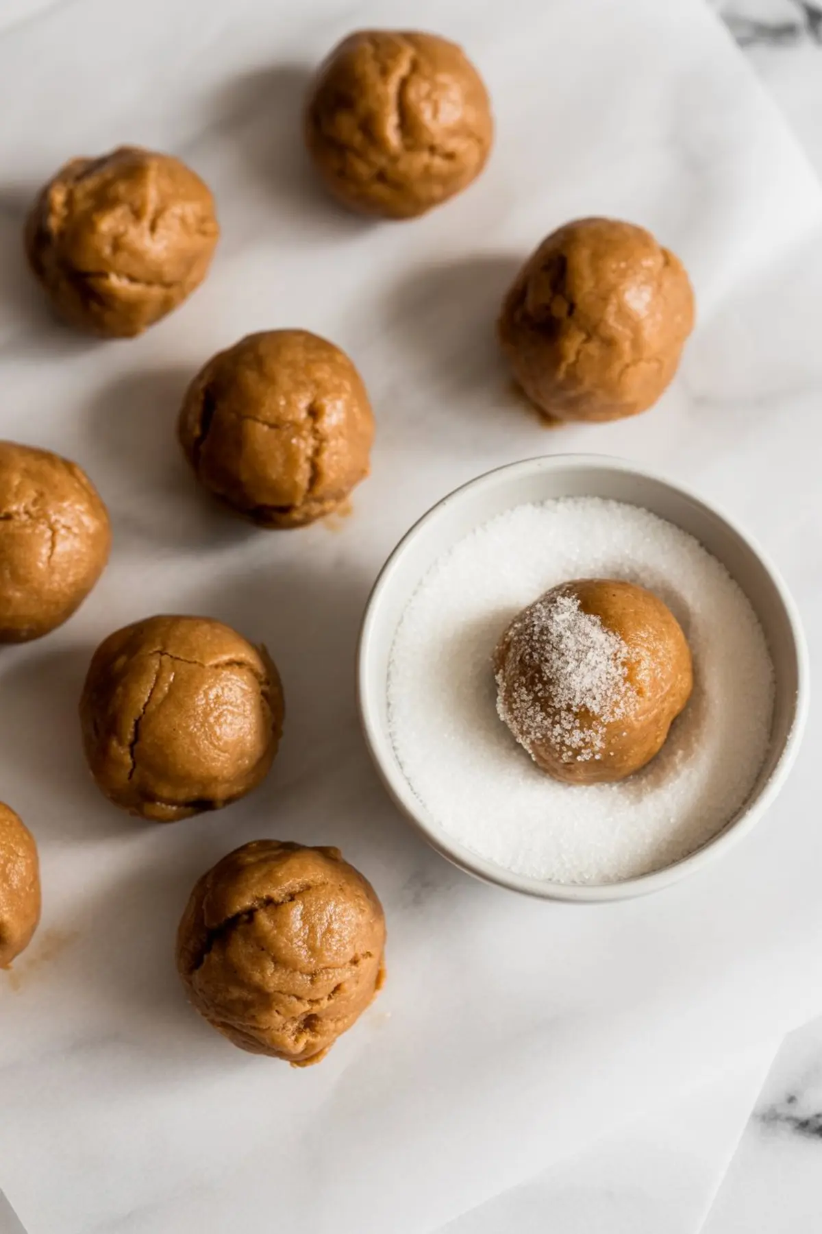 Molasses cookie dough balls on parchment paper, with one dough ball being rolled in granulated sugar in a white bowl.
