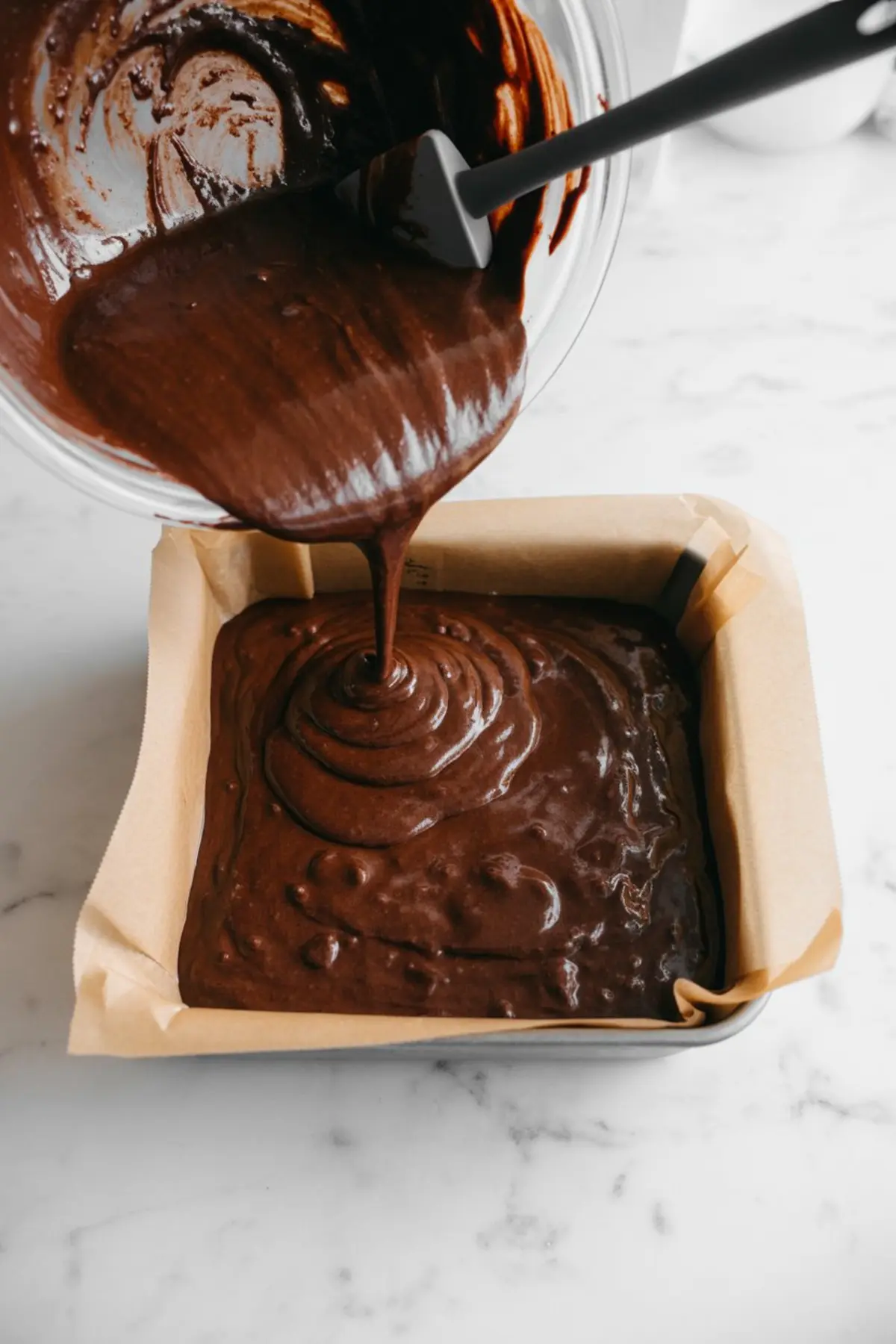 Brownie batter being poured from a glass mixing bowl into a parchment-lined square baking pan, showing a thick and glossy chocolate texture mid-pour.