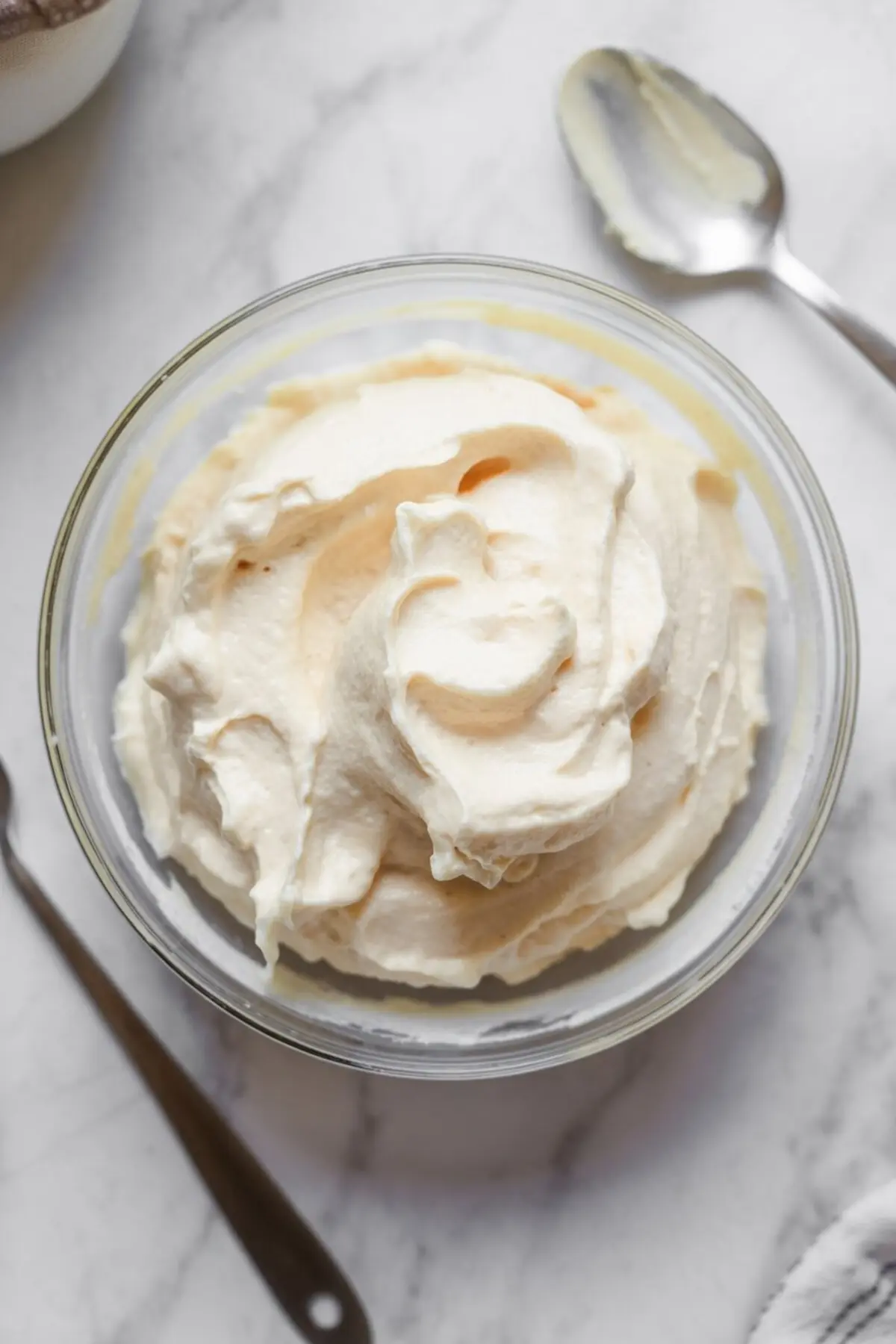 Smooth whipped vanilla cream filling in a glass bowl with a spoon on the side, ready for assembling oatmeal cream pie cookies.
