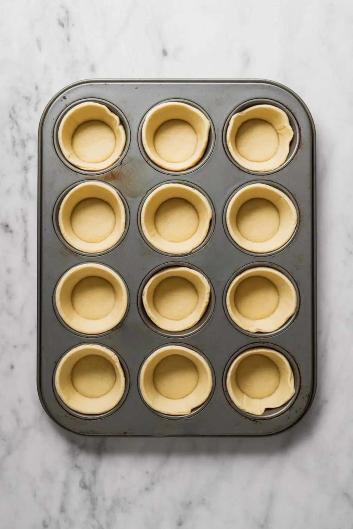 Muffin tin filled with raw puff pastry dough shells arranged in twelve circular molds, prepared for baking pastel de nata custard tarts.