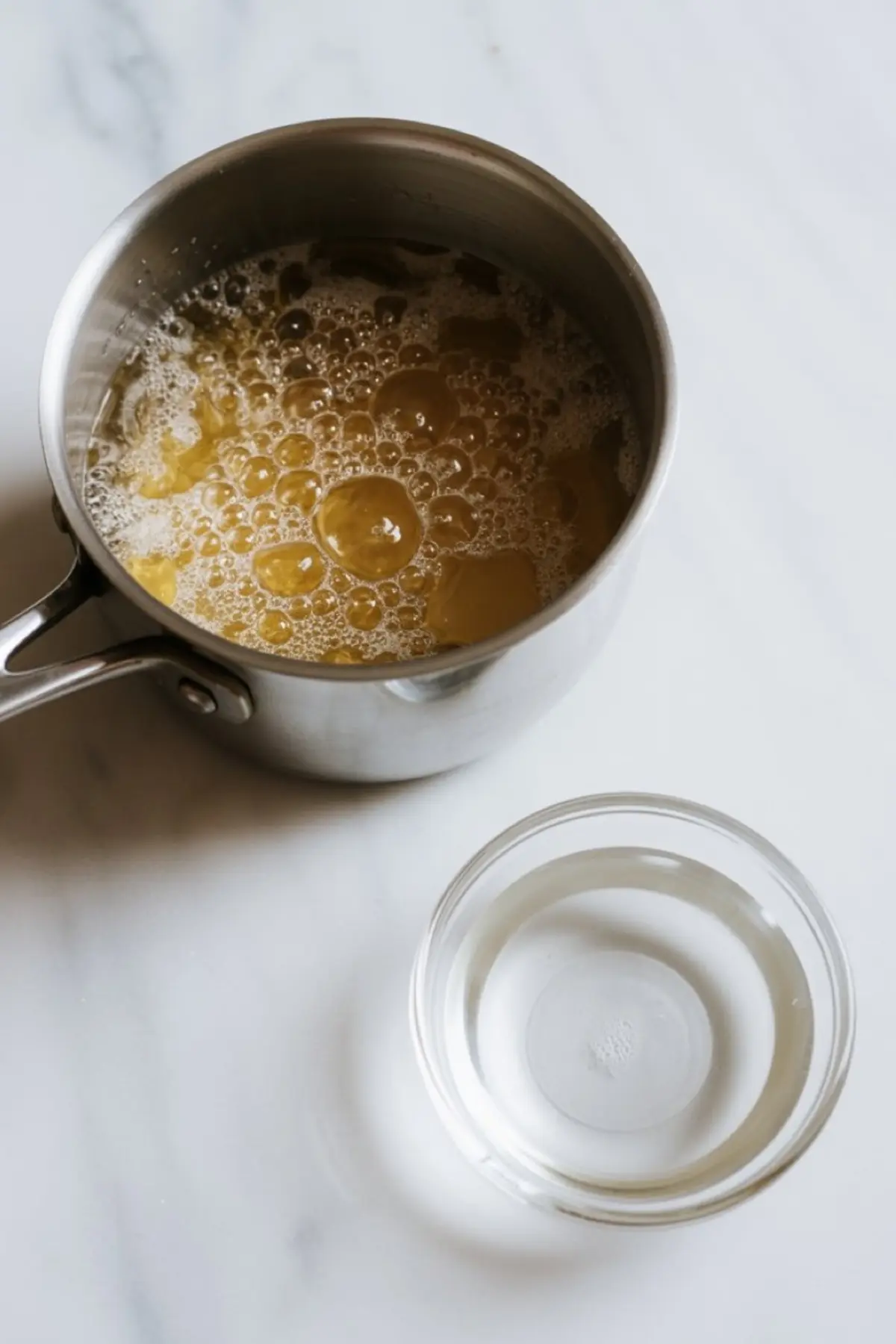 Stainless steel saucepan filled with bubbling hot syrup and a small glass bowl of clear liquid, both placed on a white countertop.