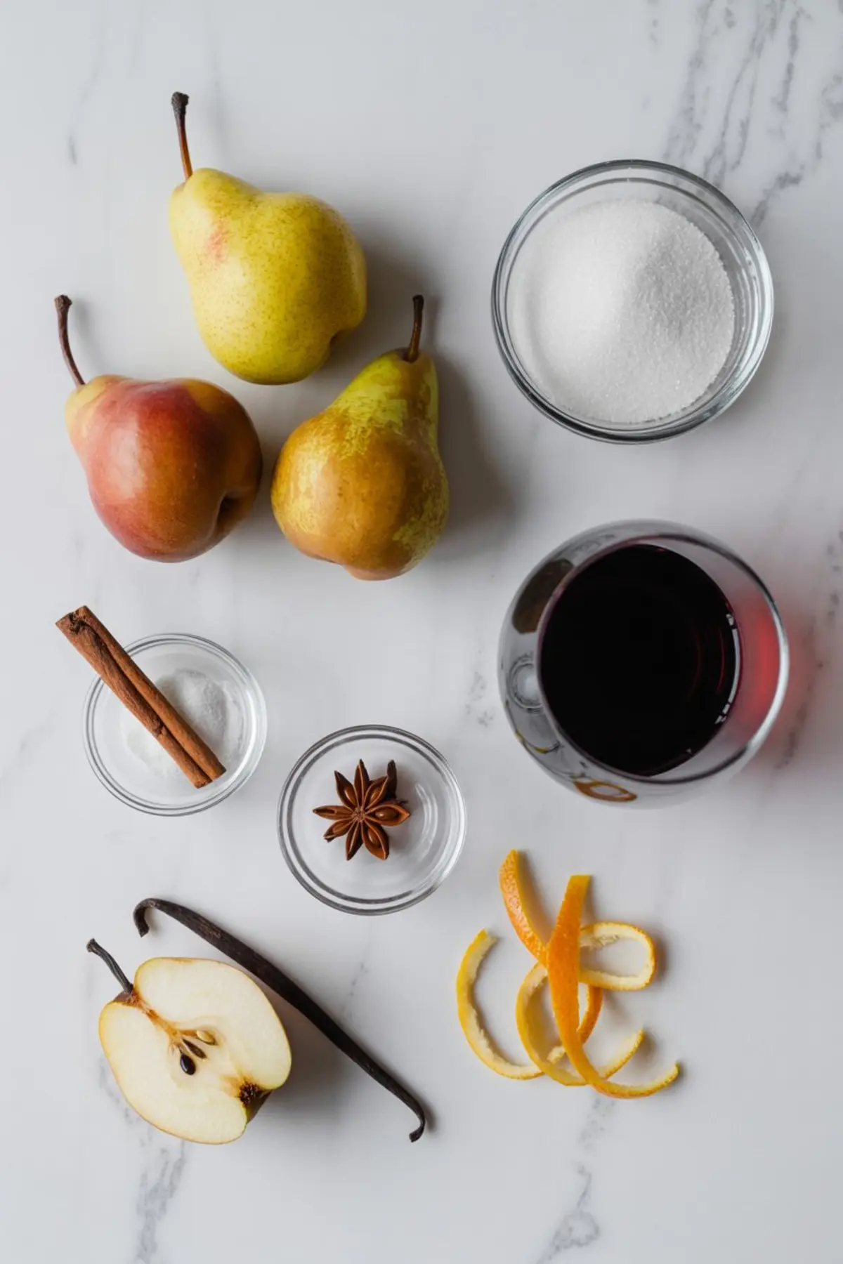Flat lay of ingredients for poached pears including fresh pears, granulated sugar, red wine, cinnamon sticks, star anise, vanilla bean, and orange peels on a marble countertop.
